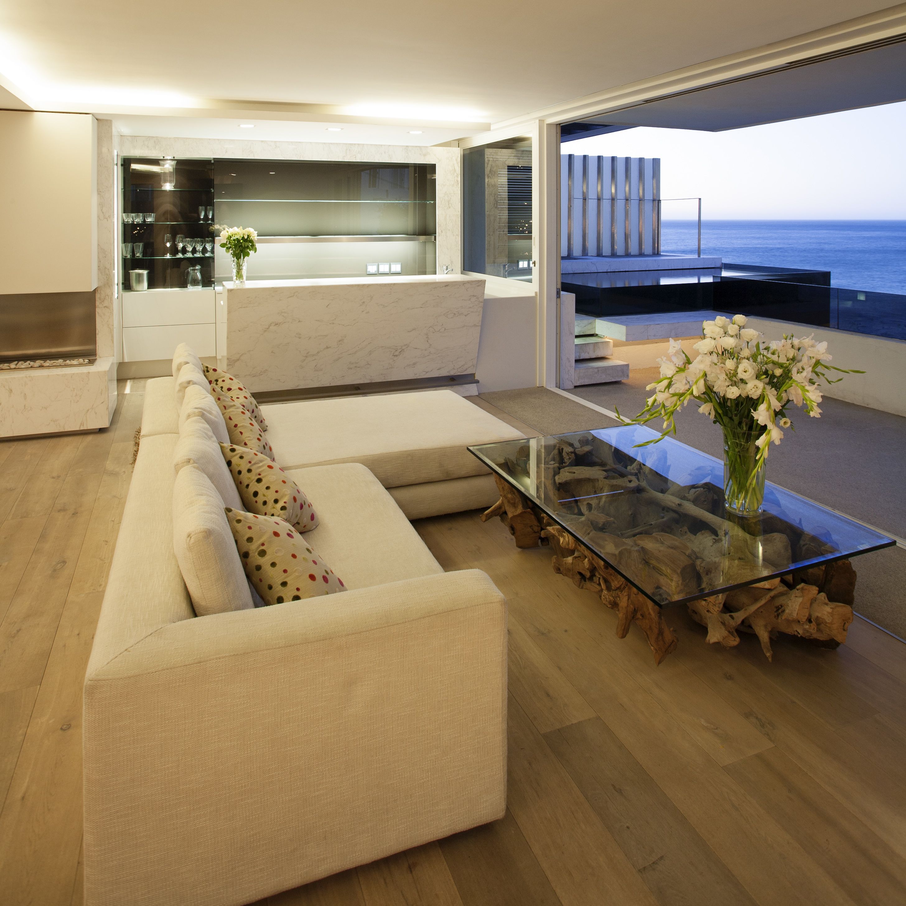 Una sala de estar moderna con una mesa de centro de vidrio, un sofá color beige con cojines estampados y vista al océano a través de ventanas amplias.