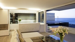 A modern living room with a glass coffee table, a beige sofa with patterned cushions, and a view of the ocean through large windows. - Ebb Tide (Cape Town)