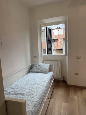 Ein kleines Schlafzimmer mit einem Einzelbett, einem Fenster mit RolllÀden, einem Heizkörper und Blick auf roten ZiegeldÀcher.