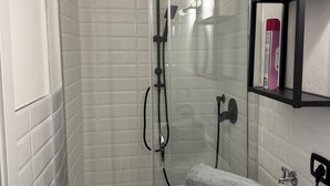A glass-enclosed shower with a white tiled wall, a towel on a white box, and a black shelf with items. -  Artdeco Luxury suite Pescarenico 2 (Lecco)