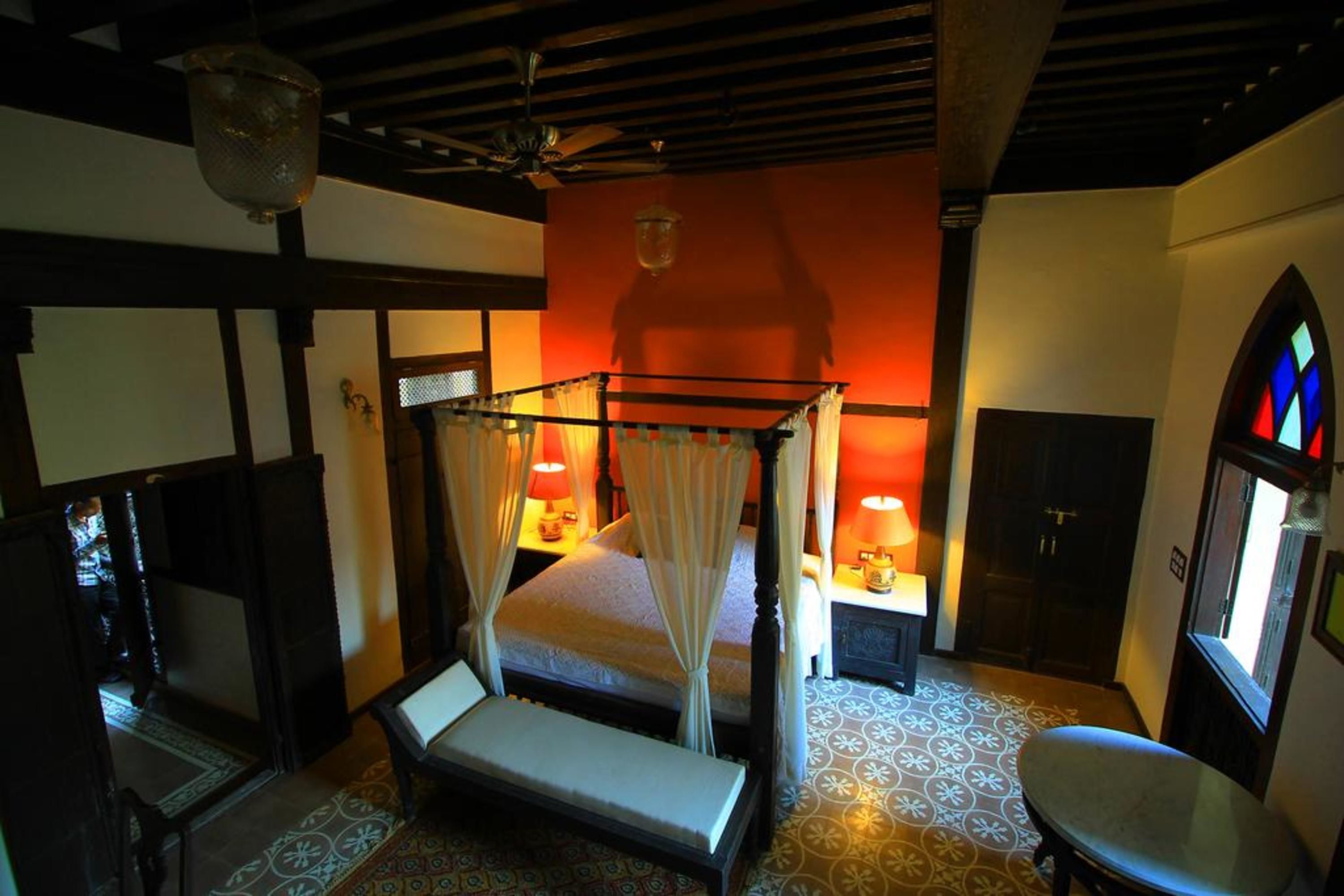 A four-poster bed with a canopy, bedside tables with lamps, a chair, and a window with stained glass.