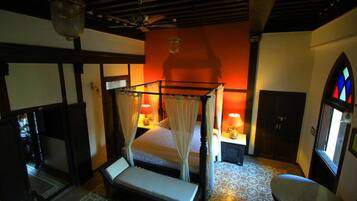 A four-poster bed with a canopy, bedside tables with lamps, a chair, and a window with stained glass.