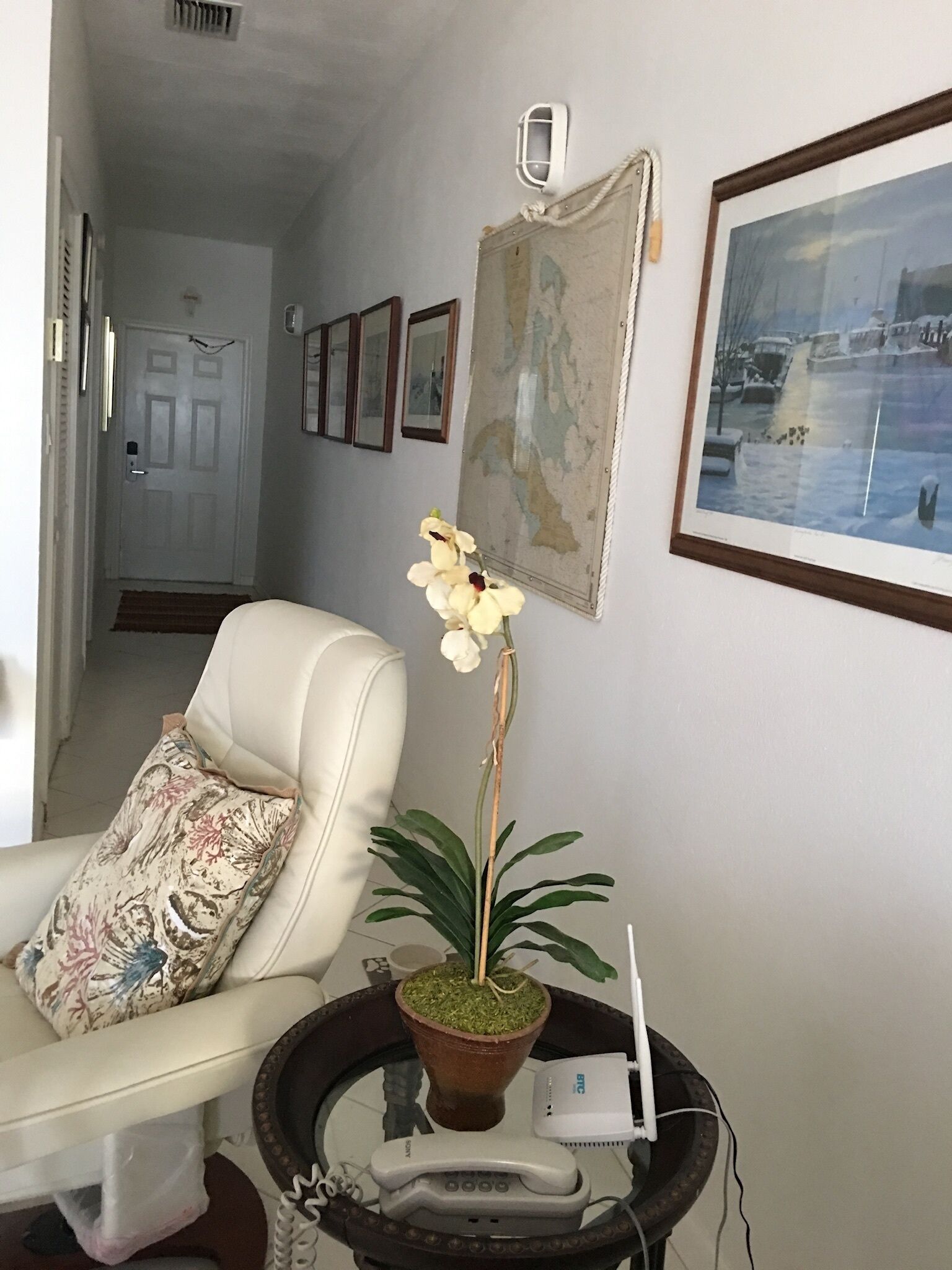 Una sala de estar con un sofá blanco, un cojín estampado, una planta en maceta, un cuadro enmarcado y una mesita con un teléfono y un router.