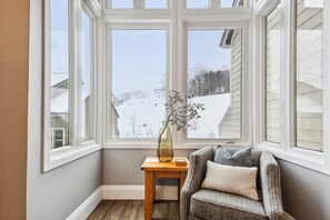 Een hoekappartement met een groot raam, een klein houten tafeltje en een fauteuil met kussens.