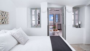 Un dormitorio con una cama, una mesita con un florero y vista a un comedor por una puerta abierta.