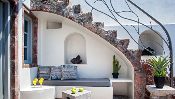 Un espacio al aire libre con luz solar, que incluye una banca de piedra, una mesa pequeña con bebidas y plantas en macetas.