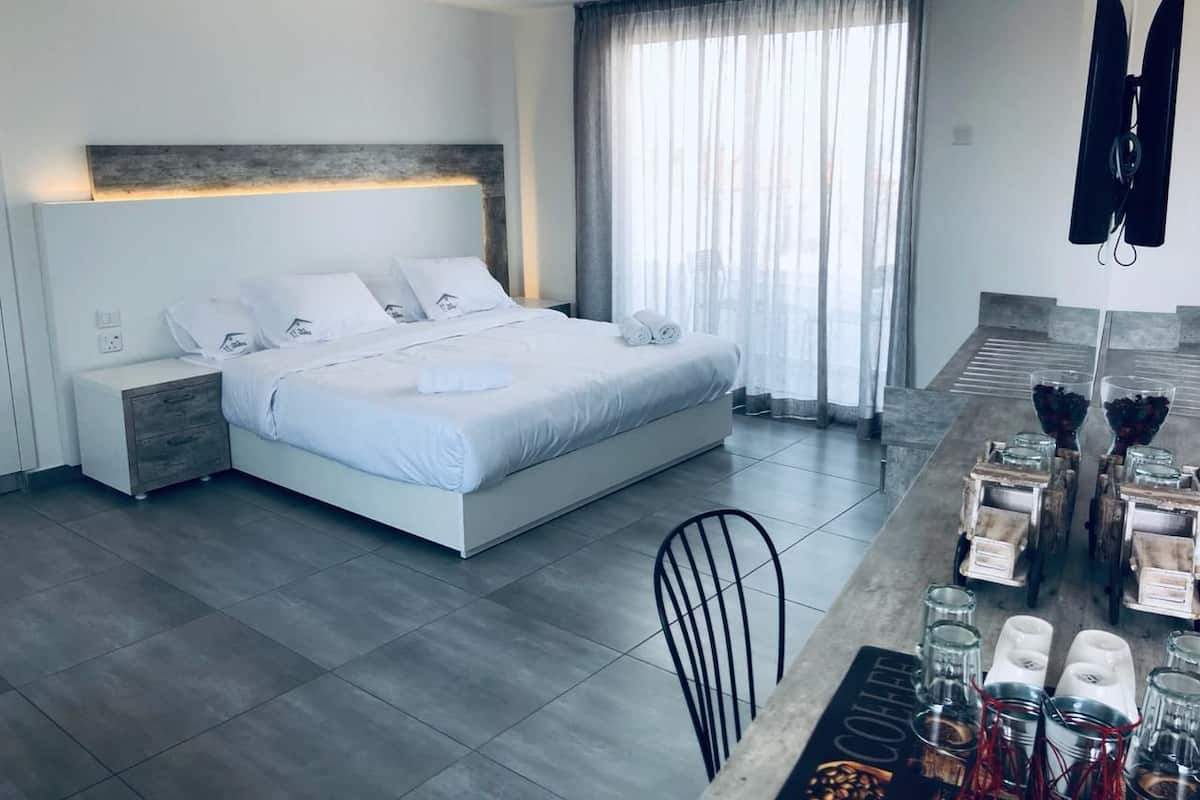 Quarto de hotel moderno com uma cama grande, uma mesinha de cabeceira, uma televisão e um espaço para café da manhã com copos e tigelas.