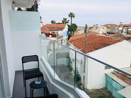 Un balcón con dos sillas negras y una mesita, con vistas a una zona residencial de techos rojos y palmeras.