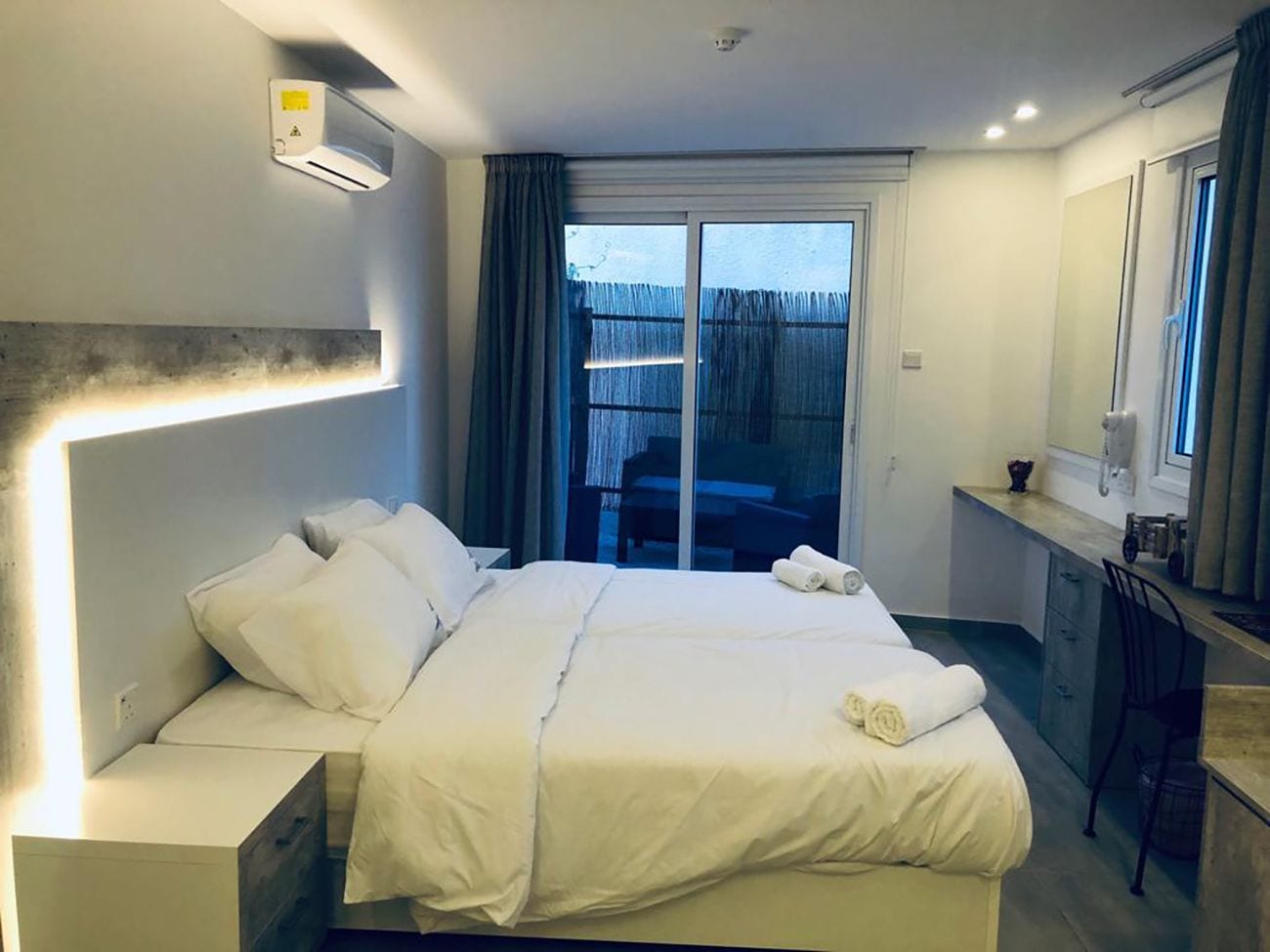 A hotel room with a large bed, a desk, a chair, and a sliding glass door leading to an outdoor area.