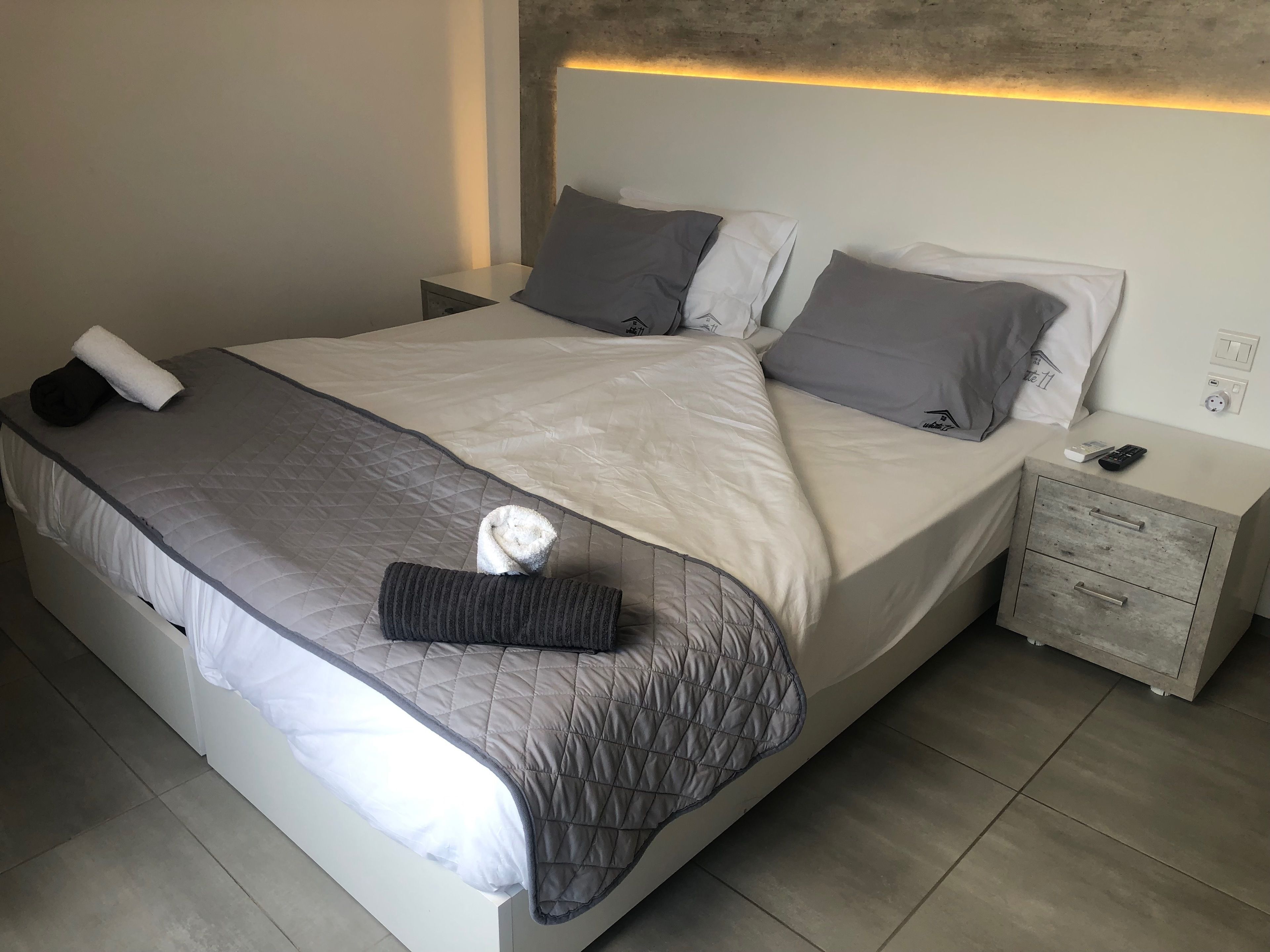 A neatly made bed with a quilted cover, pillows, and a folded towel. There is a nightstand with a remote control and a power outlet.