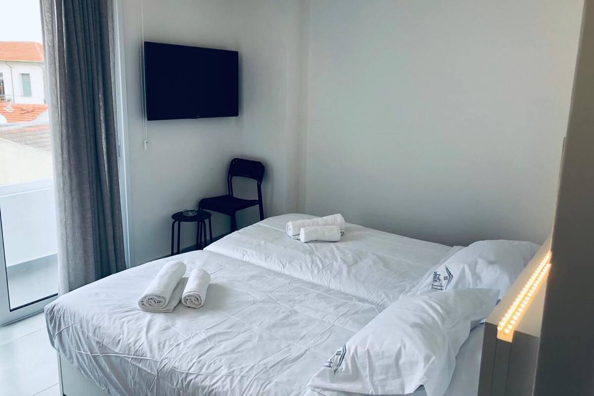 Quarto de hotel com uma cama, televisão de tela plana na parede, uma cadeira e uma janela com cortinas.