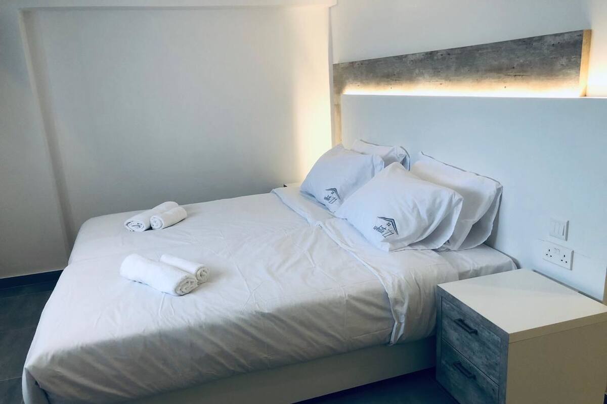 Cama bem arrumada com roupa de cama branca e toalhas dobradas, mesa de cabeceira com gavetas e luminária fixa na parede.