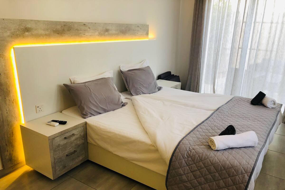 Quarto de hotel moderno com uma cama grande, mesas de cabeceira, um criado-mudo e cabeceira com iluminação integrada.
