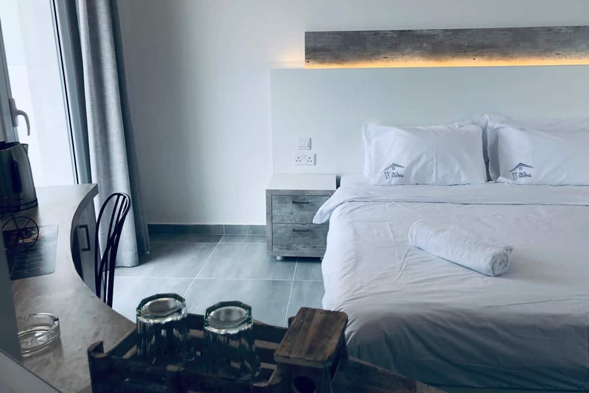 Quarto de hotel com cama bem arrumada, mesa de cabeceira com um copo, uma cadeira e um banquinho de madeira.