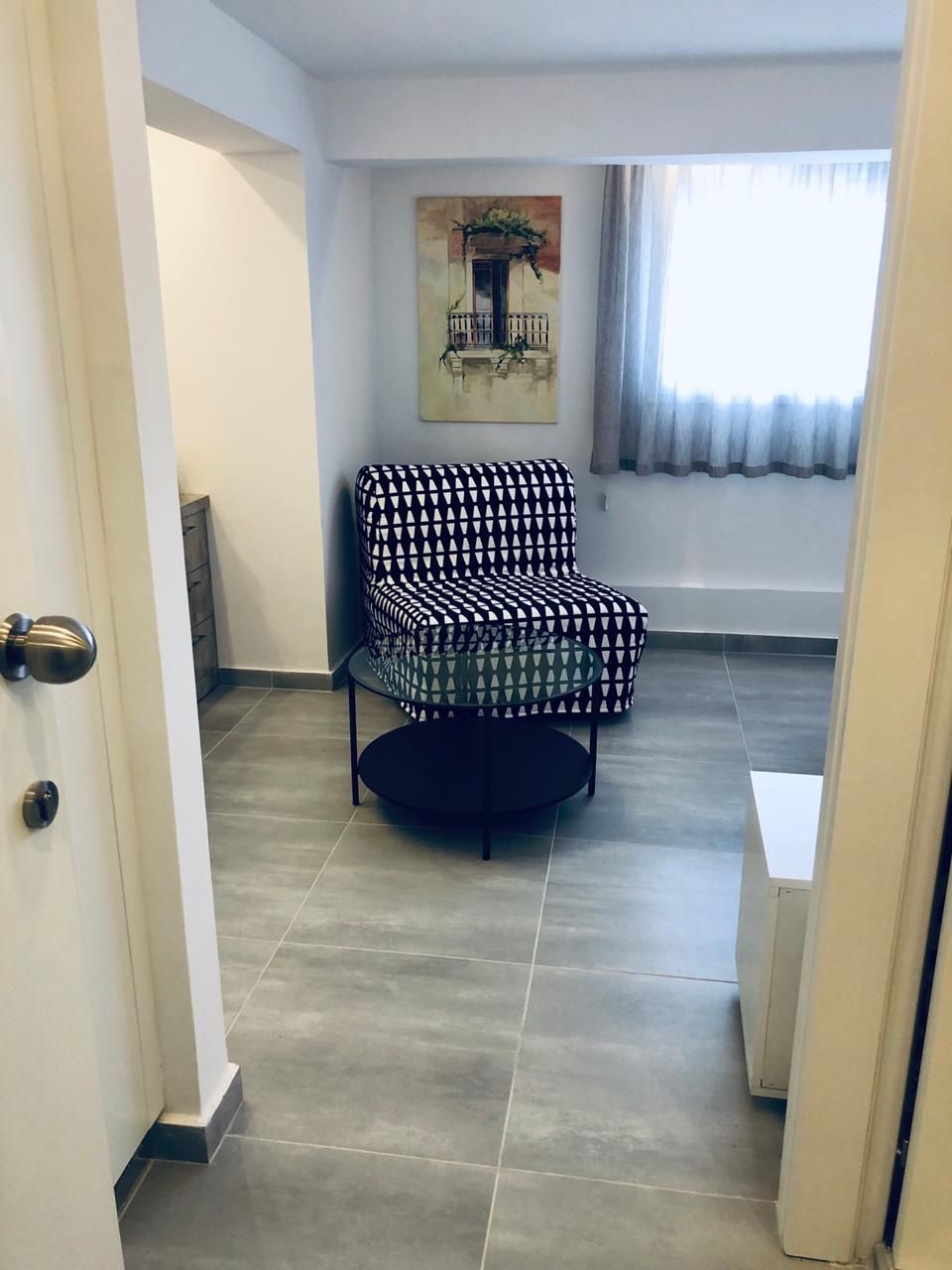A room with a patterned armchair, a painting on the wall, and a window with curtains.