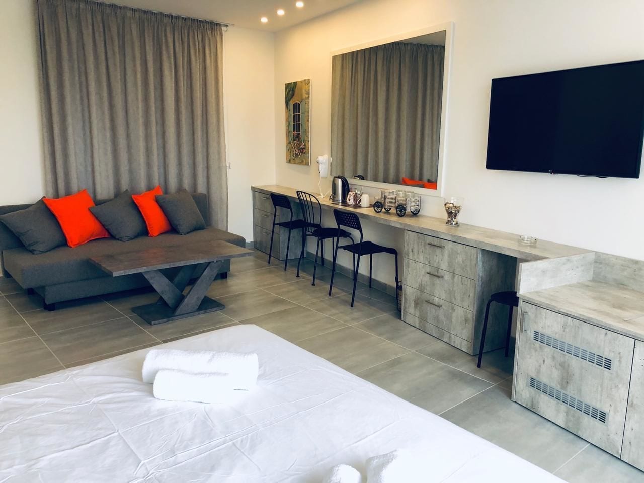 Ein modernes Hotelzimmer mit einem Bett, einer Couch mit roten Kissen, einer Essecke mit Stühlen, einer Küchenzeile und einem an der Wand befestigten Flachbildfernseher.