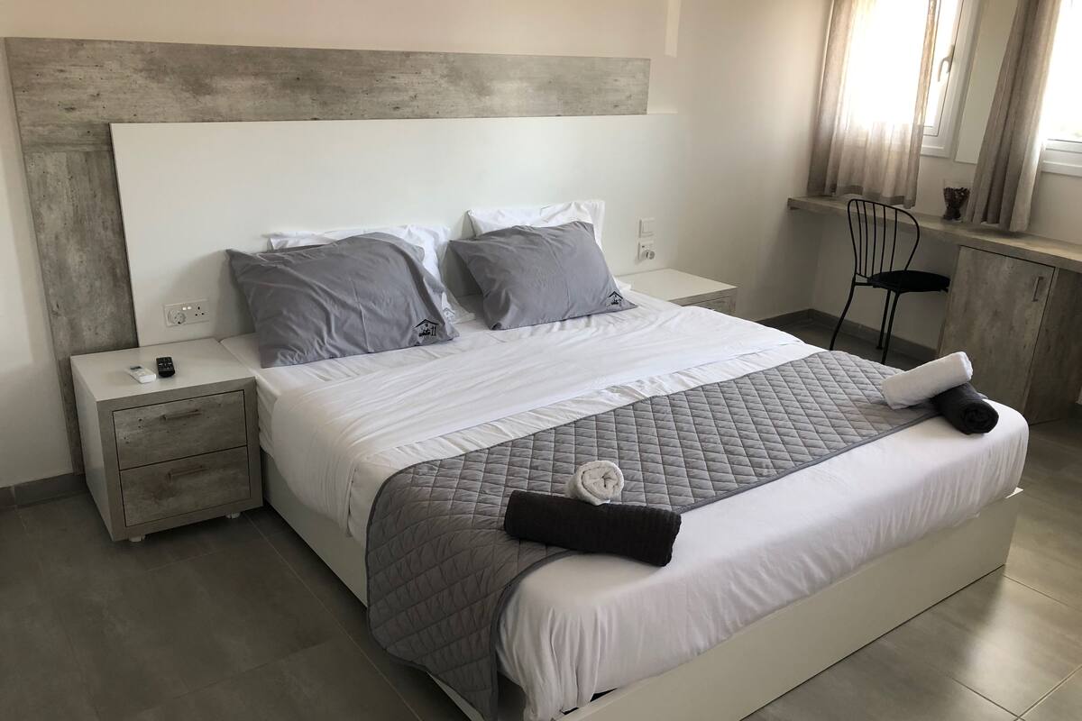 Quarto moderno com cama grande, roupa de cama cinza, cabeceira de madeira, mesa de cabeceira com controle remoto e janela com cortinas.