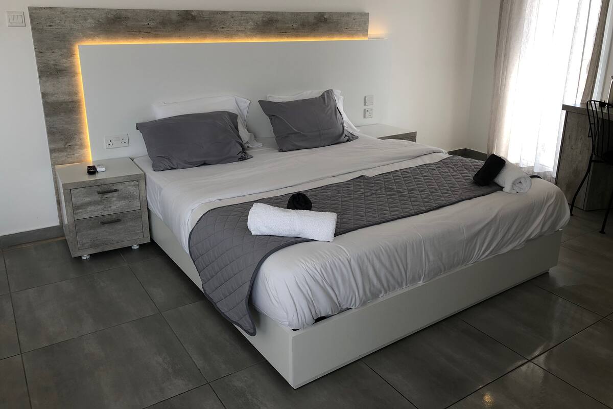 Quarto moderno com uma cama grande, roupa de cama cinza, uma mesa de cabeceira com controle remoto e um criado-mudo.