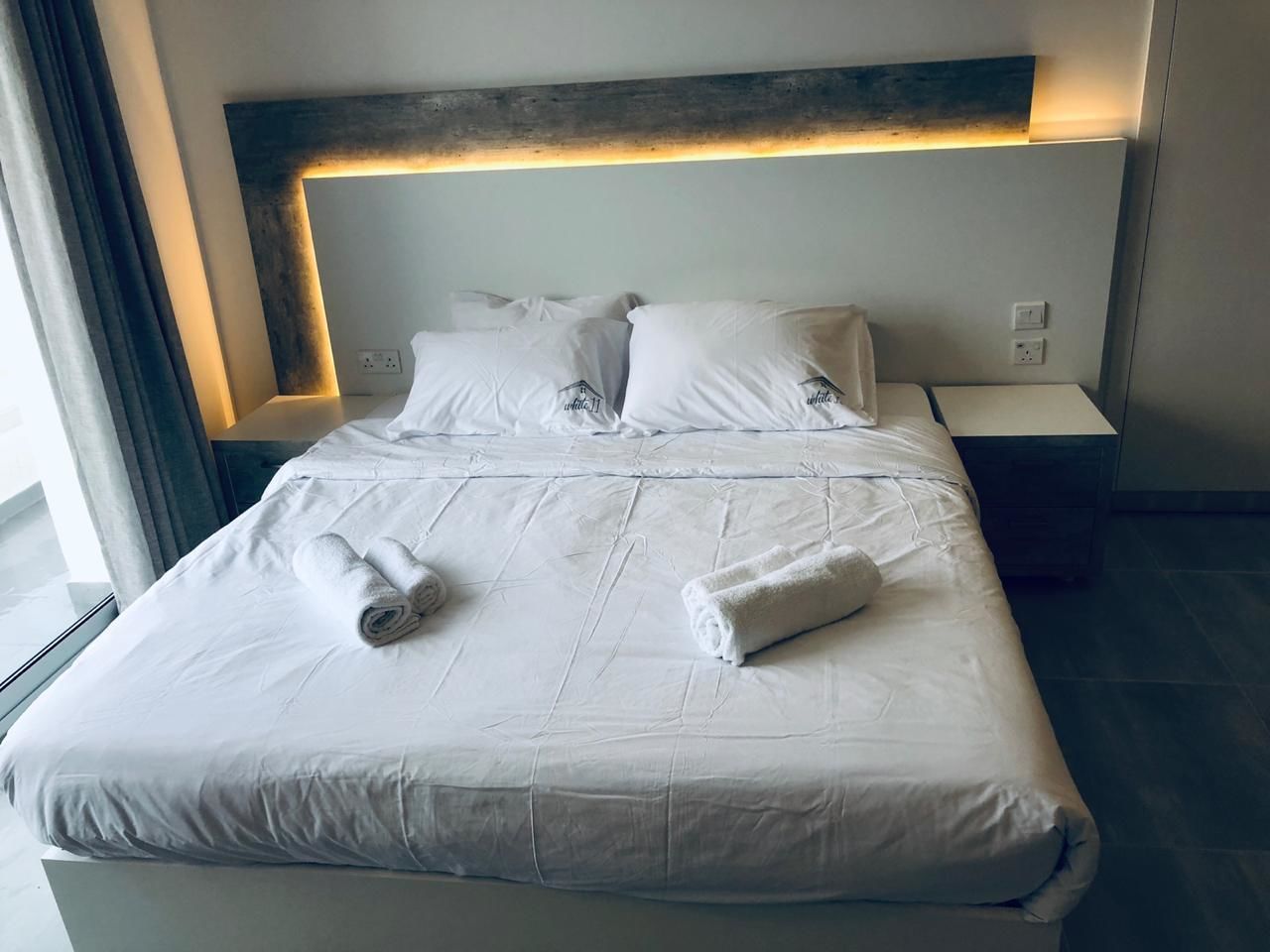 A neatly made bed with white linens and two rolled towels, a wooden headboard with integrated lighting, and a small bedside table with a power outlet.