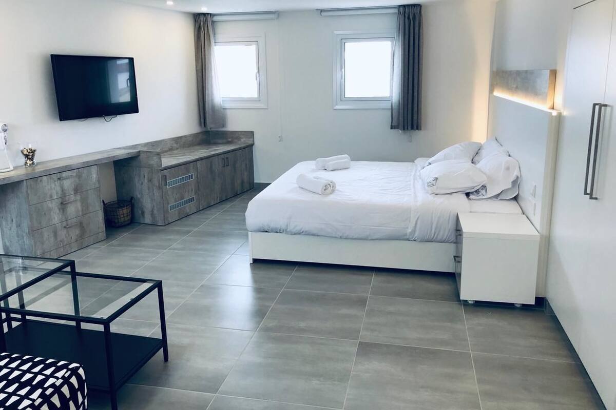 Quarto de hotel moderno com cama, mesas de cabeceira, televisão, pequena cozinha e área de estar.