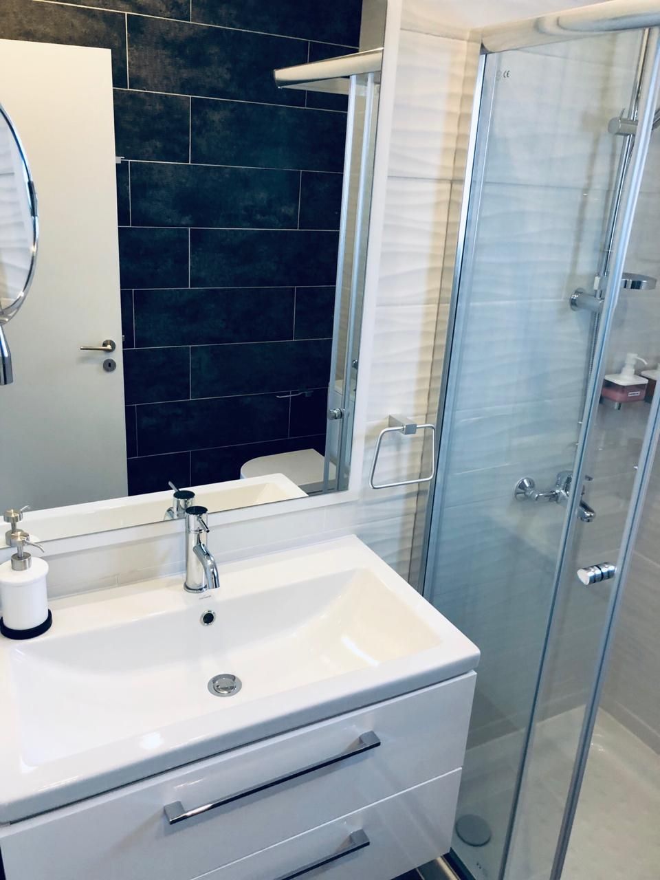 Un baño con lavamanos blanco, un espejo, cabina de ducha de vidrio y azulejos azul oscuro.