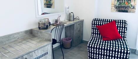 A compact room with a kitchenette, a chair, a red pillow, and two paintings on the walls.