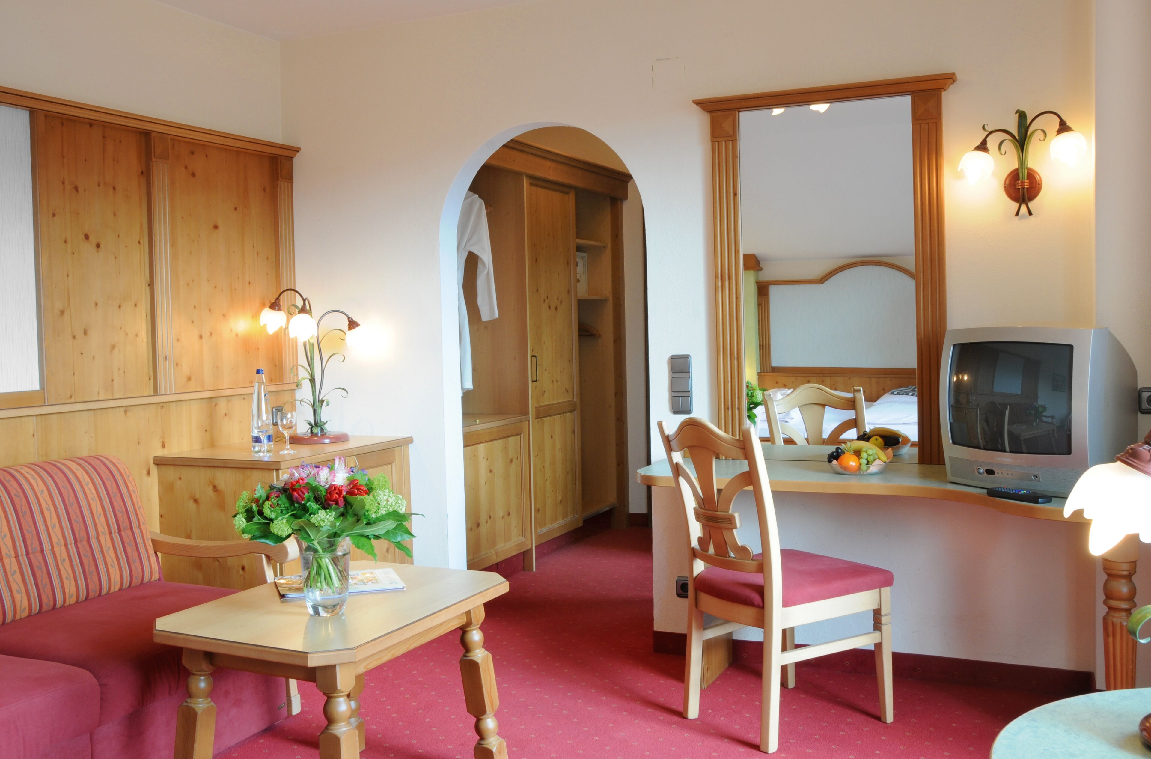 A cozy room with a wooden sofa, a dining table with a vase of flowers, a wooden chair, a television, and a mirror.