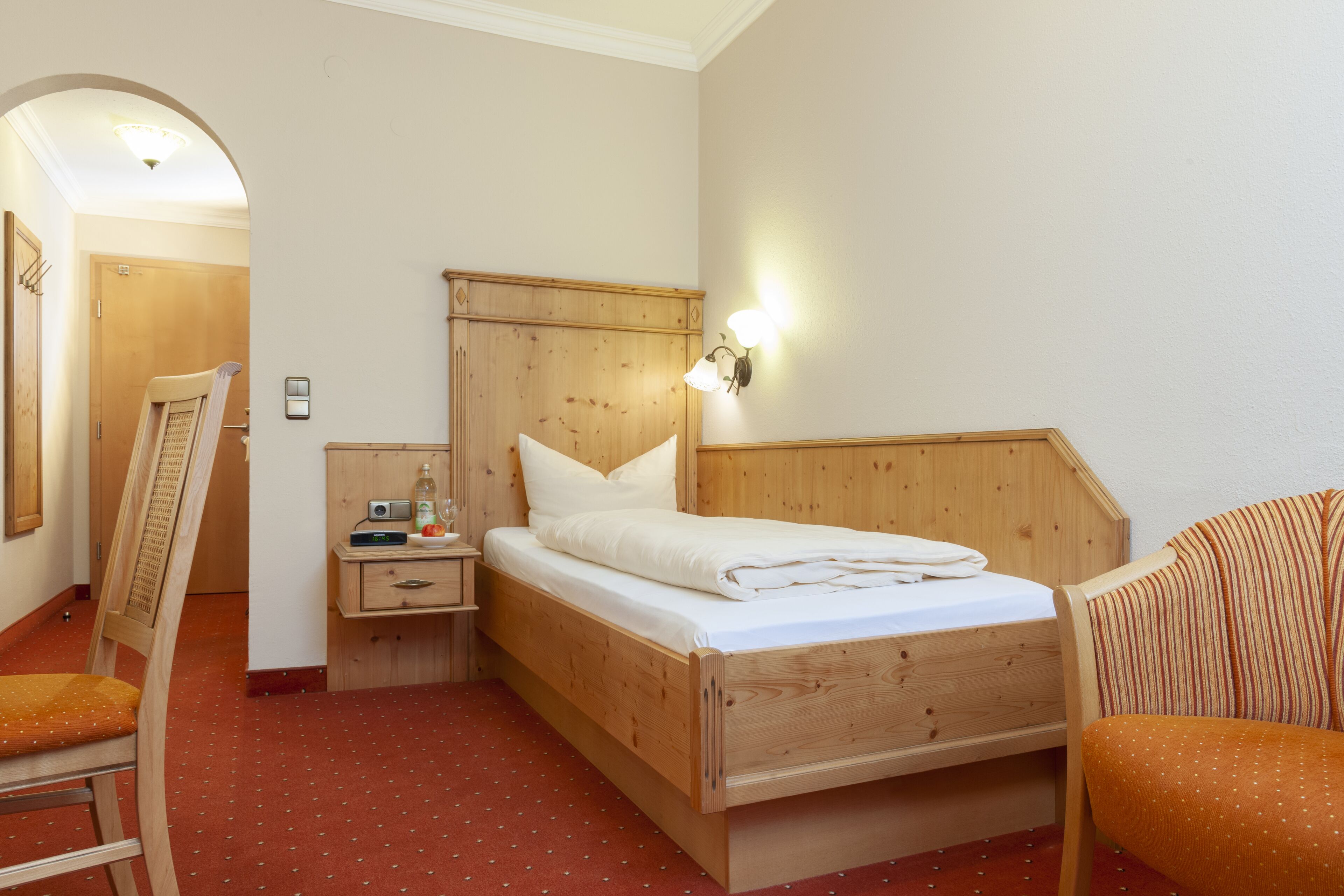 A wooden bed with a headboard, a nightstand with a radio, a chair, and a wall-mounted light fixture.