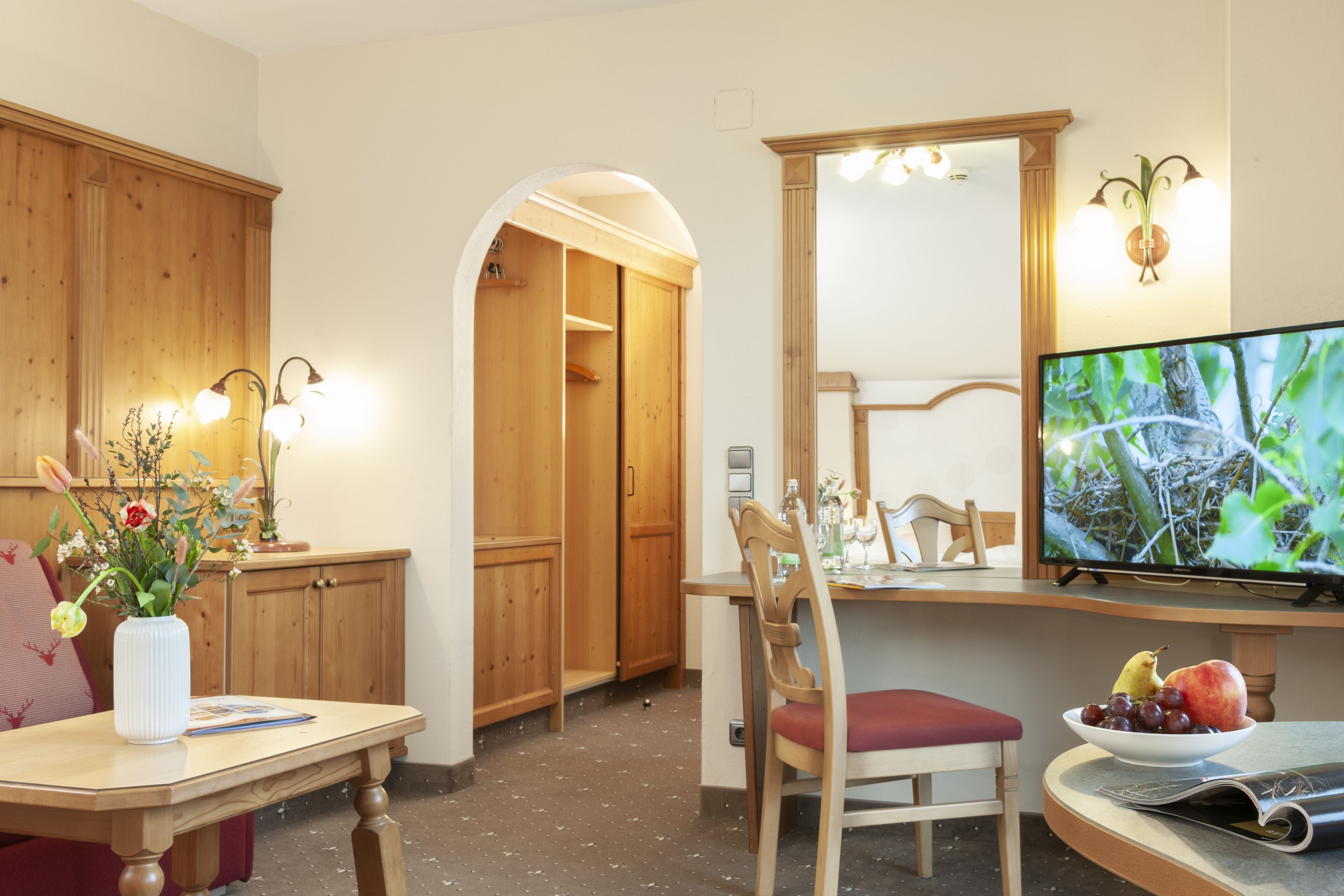 Una habitación con armarios de madera, una mesa de comedor con un florero y un televisor sobre un mueble, además de un frutero con fruta.