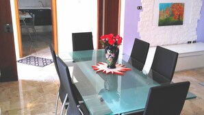 Un área de comedor con una mesa de vidrio, sillas negras, un jarrón con flores rojas, un muro de ladrillo blanco y un cuadro.