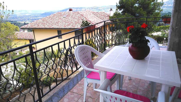 Un balcón con una mesa y sillas blancas, una planta en maceta y vistas de una casa con tejado rojo y un paisaje lejano.