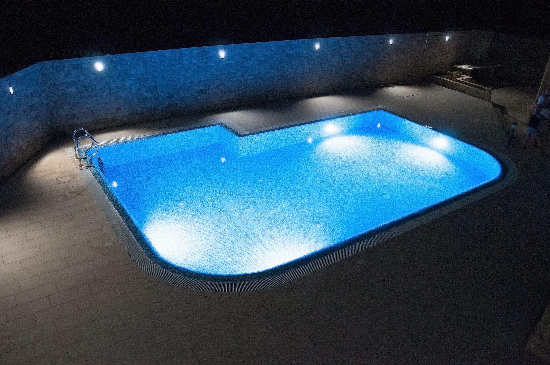 A rectangular swimming pool with a textured surface, surrounded by a brick wall and illuminated by several lights.