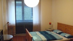 Une chambre à coucher avec un lit, une petite table, une chaise, une lampe et une vue sur l’extérieur, visible à travers une porte.