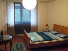 A bedroom with a bed, a small table, a chair, a lamp, and a view through a door.