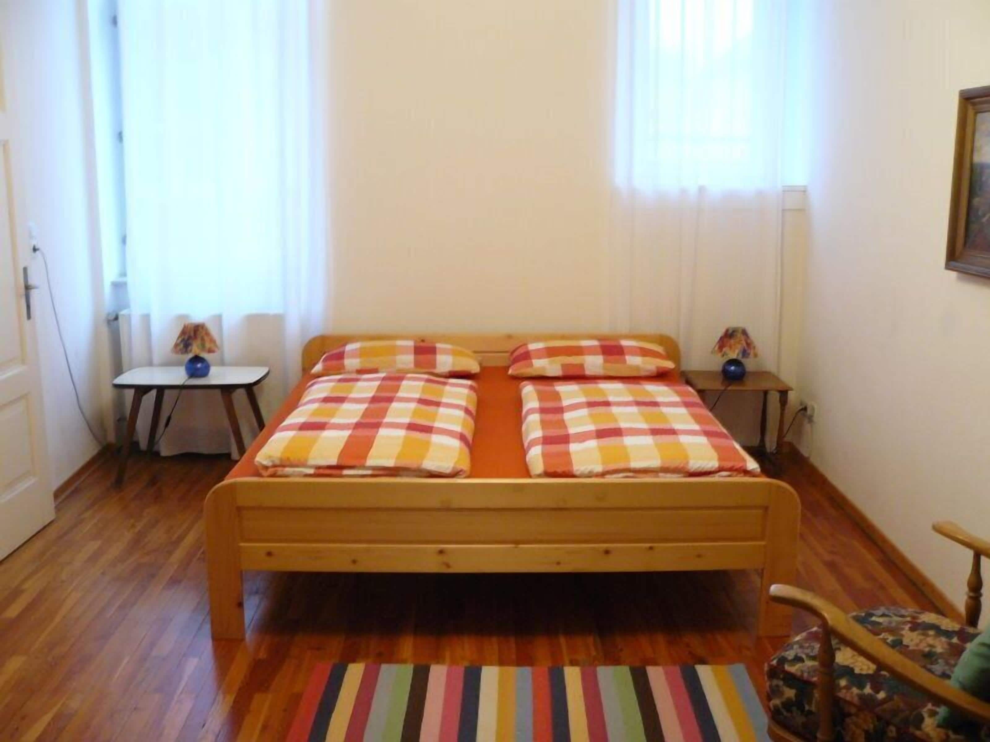 A single bed with a checkered bedspread, a small table with a lamp, a chair, and a striped rug on a wooden floor.
