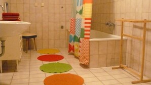 Une salle de bain avec un lavabo, des toilettes et une baignoire, tous de couleur blanche. Le sol est recouvert de tapis colorés et il y a un rideau de douche orné de différents motifs.