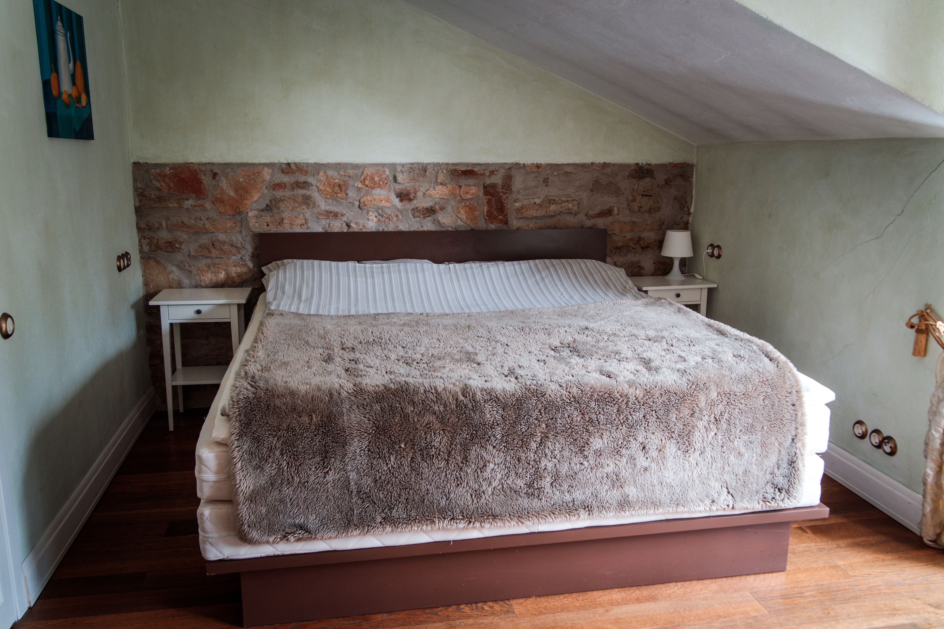 A bedroom with a stone wall, a wooden bed frame, a nightstand, and a bedside lamp.
