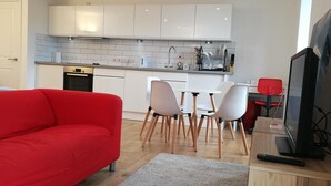 A modern kitchen with white cabinets, a dining area with white chairs, a red sofa, and a TV on a wooden stand. - Awesome City Apartment (Cambridge)