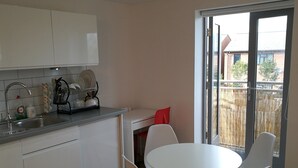 A compact kitchen with white cabinets, a sink, and a dish rack. There is a round table with four white chairs and a red pillow. A sliding glass door leads to an outdoor area with a bamboo fence. - Awesome City Apartment (Cambridge)