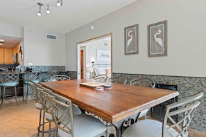 Un grand espace de salle à manger avec une table en bois et des chaises en métal, une cuisine avec un dosseret de carrelage bleu, et deux œuvres d’art encadrées accrochées au mur.