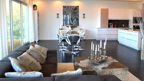 Uma sala de estar moderna com um sofá cinza escuro, uma área de jantar com mesa de vidro e uma cozinha com armários brancos.