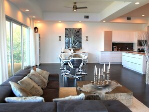 Una sala de estar moderna con un sofá gris oscuro, un comedor con mesa de vidrio y una cocina con armarios blancos.