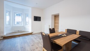 A modern dining area with a wooden table and black chairs, a flat-screen TV mounted on the wall, and large windows with white blinds. - Appartementhaus Hohe Tauern (Obervellach)