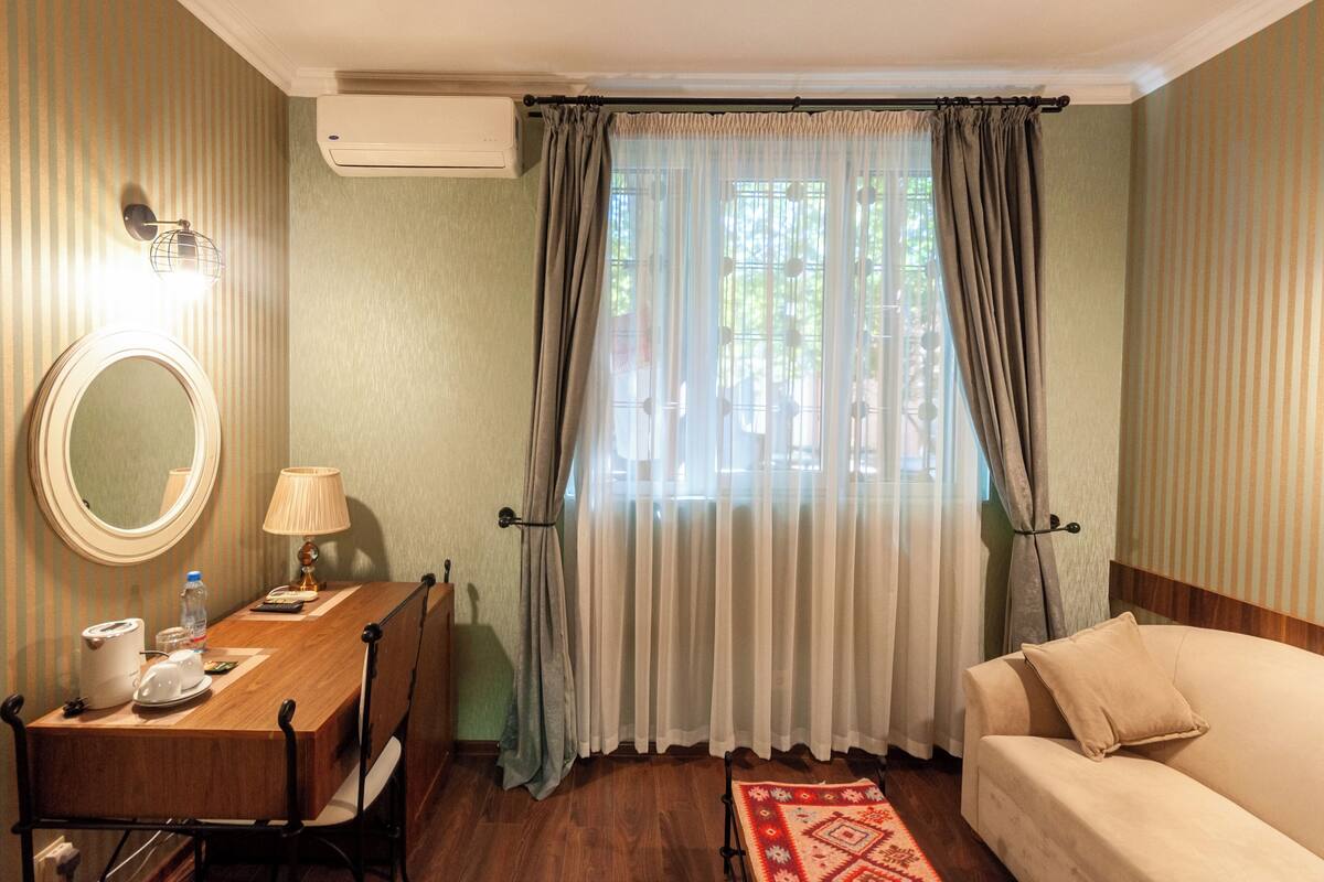 A hotel room with a wooden dining table, a round mirror, a lamp, a sofa, and a window with curtains.
