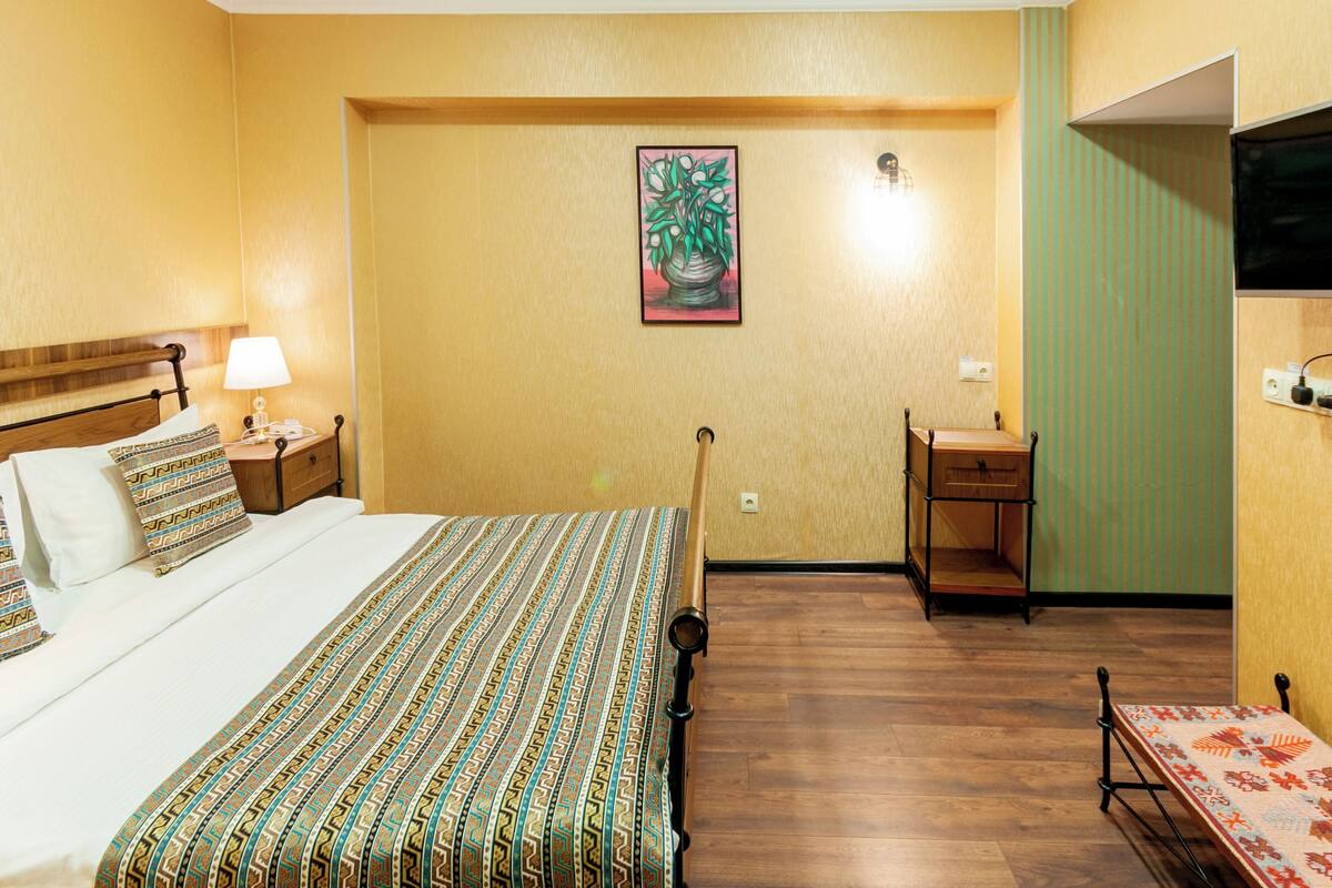 A hotel room with a bed, bedside table, lamp, desk, chair, and a painting on the wall.