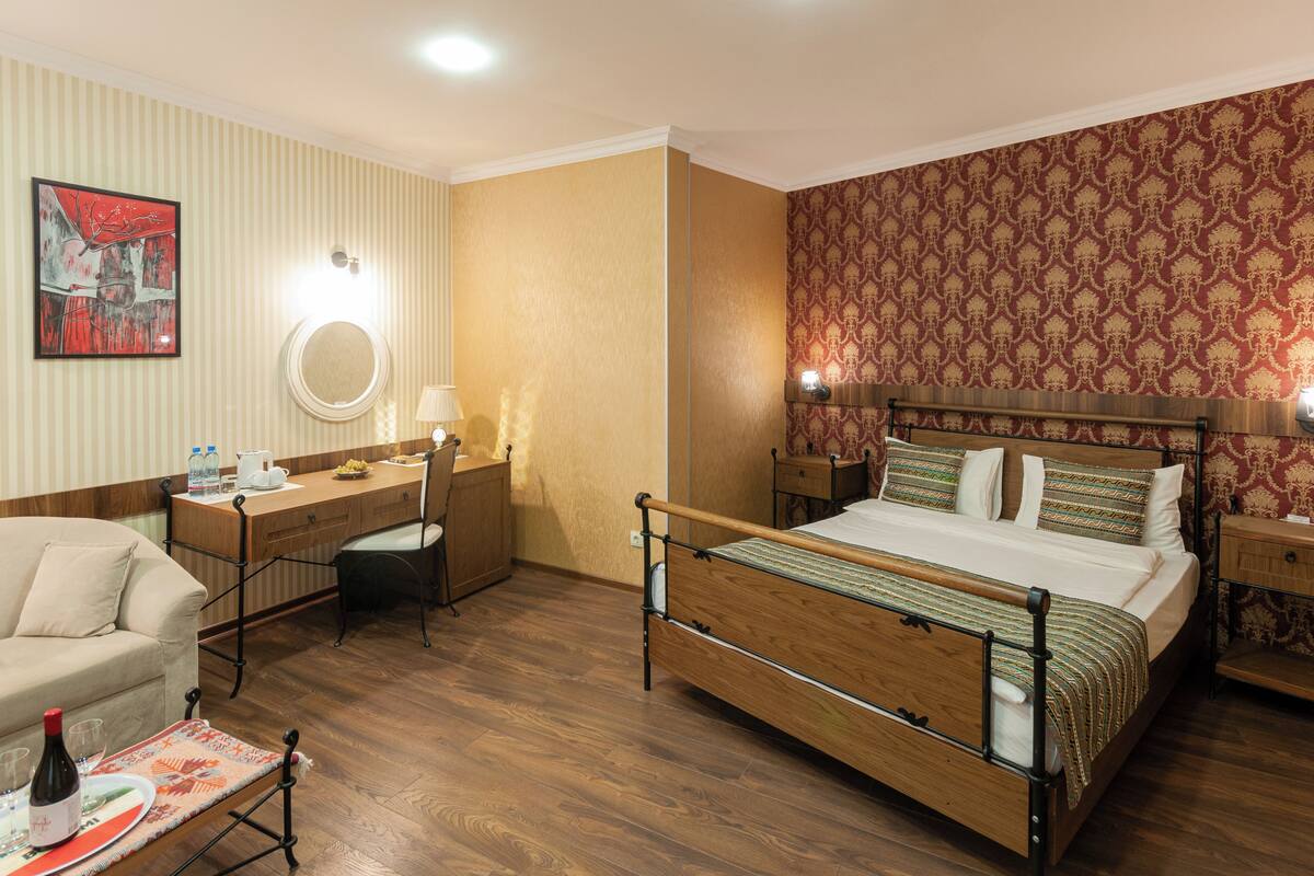 A hotel room with a wooden bed, a desk with a chair, a small table with a bottle and glasses, and a wall with a patterned wallpaper.