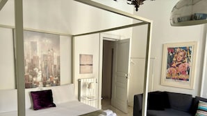 A modern bedroom with a four-poster bed, a grey sofa, and a cityscape painting. - Super central and comfortable house in historic 19th-century building. (Acireale)