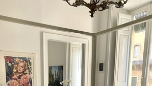 A modern living room with a dark grey sofa, a white rug, and a large mirror reflecting the room. There is a poster on the wall and a window with a view of buildings outside. - Super central and comfortable house in historic 19th-century building. (Acireale)