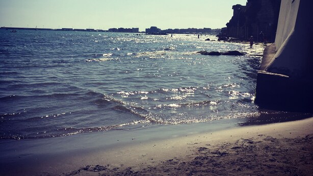 Una playa con agua cristalina, una orilla arenosa y edificios al fondo.
