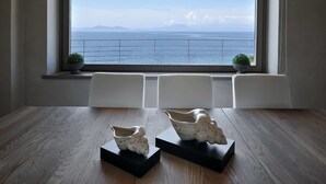 Una habitación con un gran ventanal con vista al mar, que incluye dos sillas blancas, piso de madera y dos jarrones decorativos sobre pedestales negros.